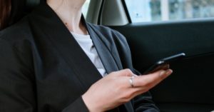 A woman with a ponytail and earrings uses a smartphone in the back of a car, focusing outside.