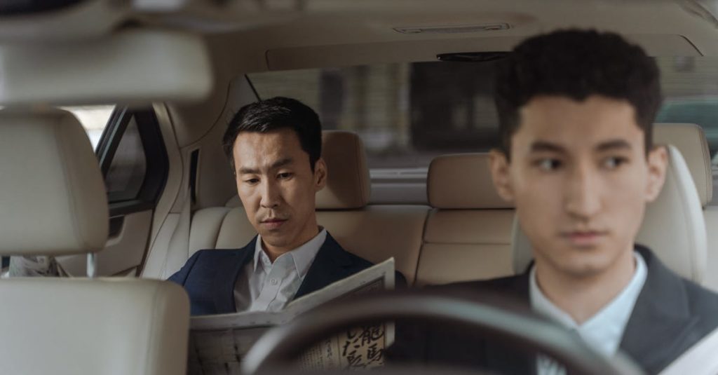 Elegant Asian businessman reading a newspaper inside a luxury car. Professional driver in focus.